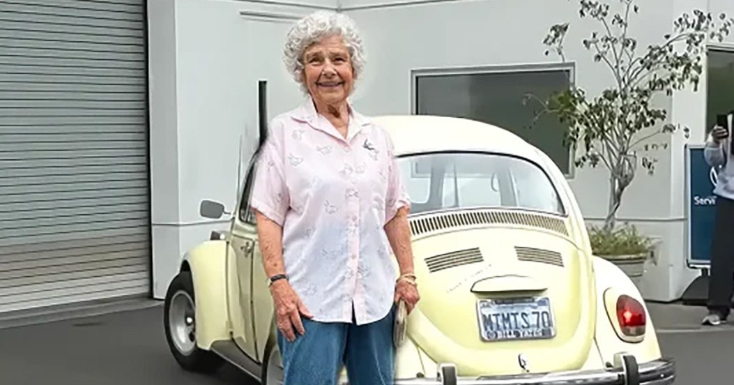 An older adult female wearing a light pink button-up shirt with small floral patterns stands next to a vintage Volkswagen Beetle at a service center, holding hands with an adult male in a light gray hoodie and black cap.