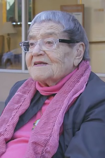 An older adult female in a wheelchair outside a building, wearing a pink sweater and glasses with a dark jacket layered under it.