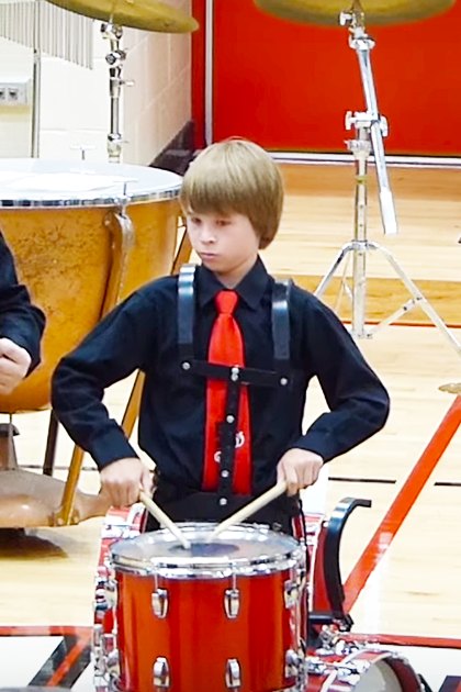Children playing drums inside a school gymnasium with wooden floors and white-red walls, wearing black shirts with red ties and black pants; drumsticks visible.