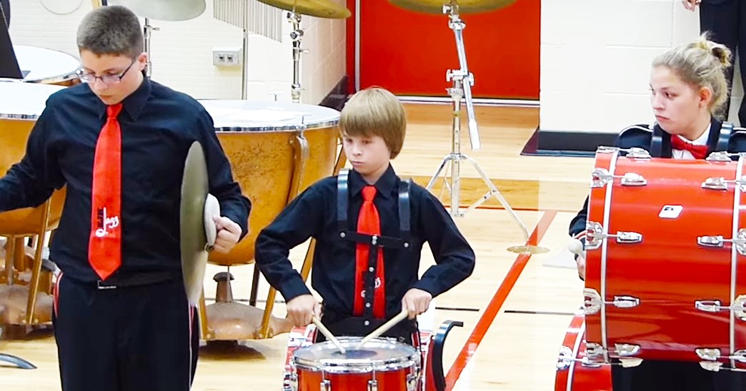 Children playing drums inside a school gymnasium with wooden floors and white-red walls, wearing black shirts with red ties and black pants; drumsticks visible.