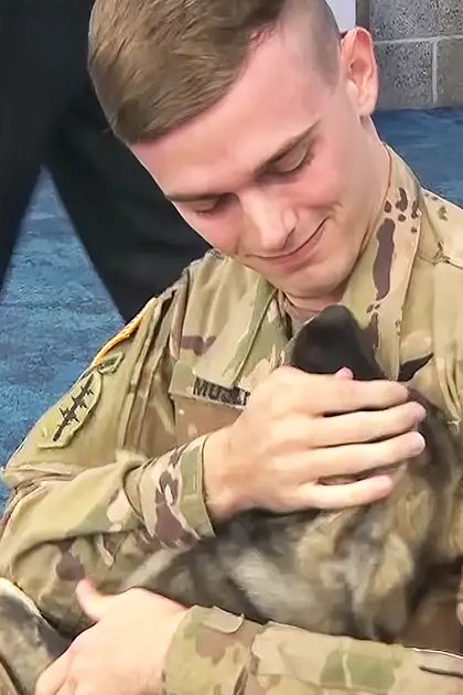 Young adult male soldier in a military uniform holding a small German Shepherd mix puppy indoors, possibly at an airport or event hall.
