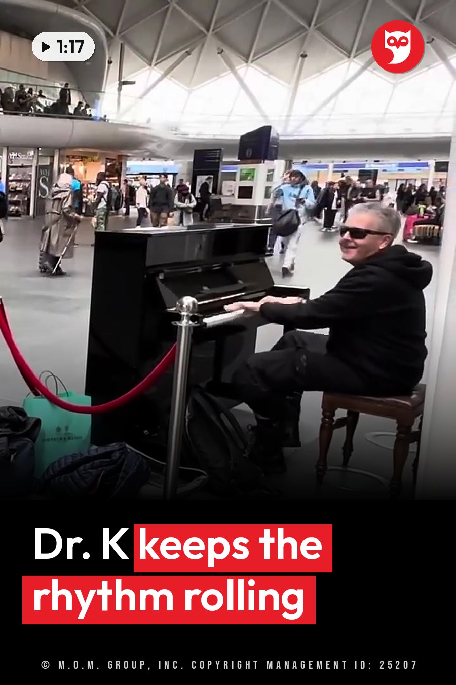 A man plays a piano in a busy indoor hall while people walk around him. A headline runs in white letters across the lower third.
