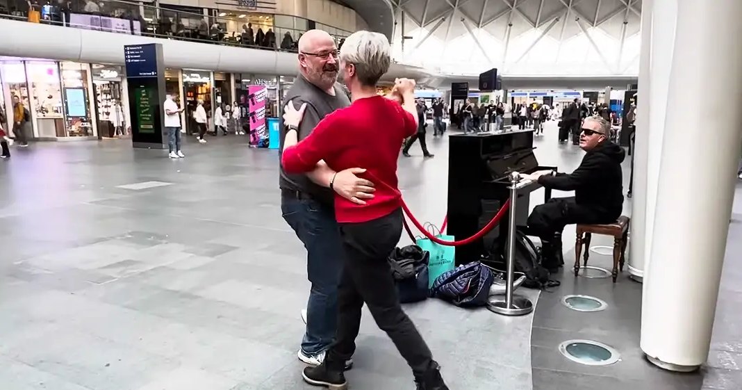 A middle-aged couple dances in a public indoor space with shopping bags visible near a seated older adult pianist, surrounded by pillars and high ceilings.