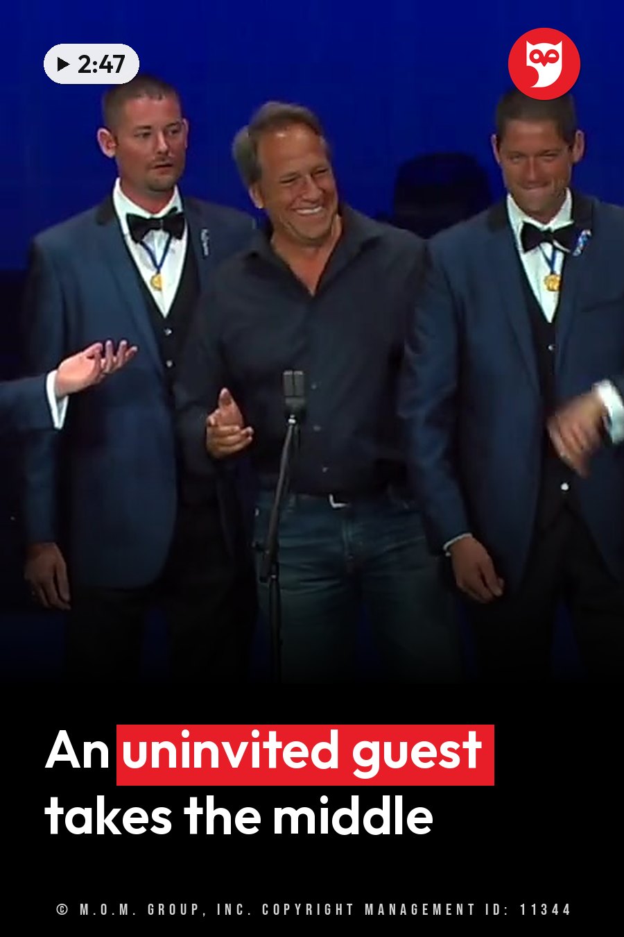 Three men stand on a stage; the man in the center laughs into a microphone, flanked by two men in suits and bow ties. A headline runs in white letters across the lower third.