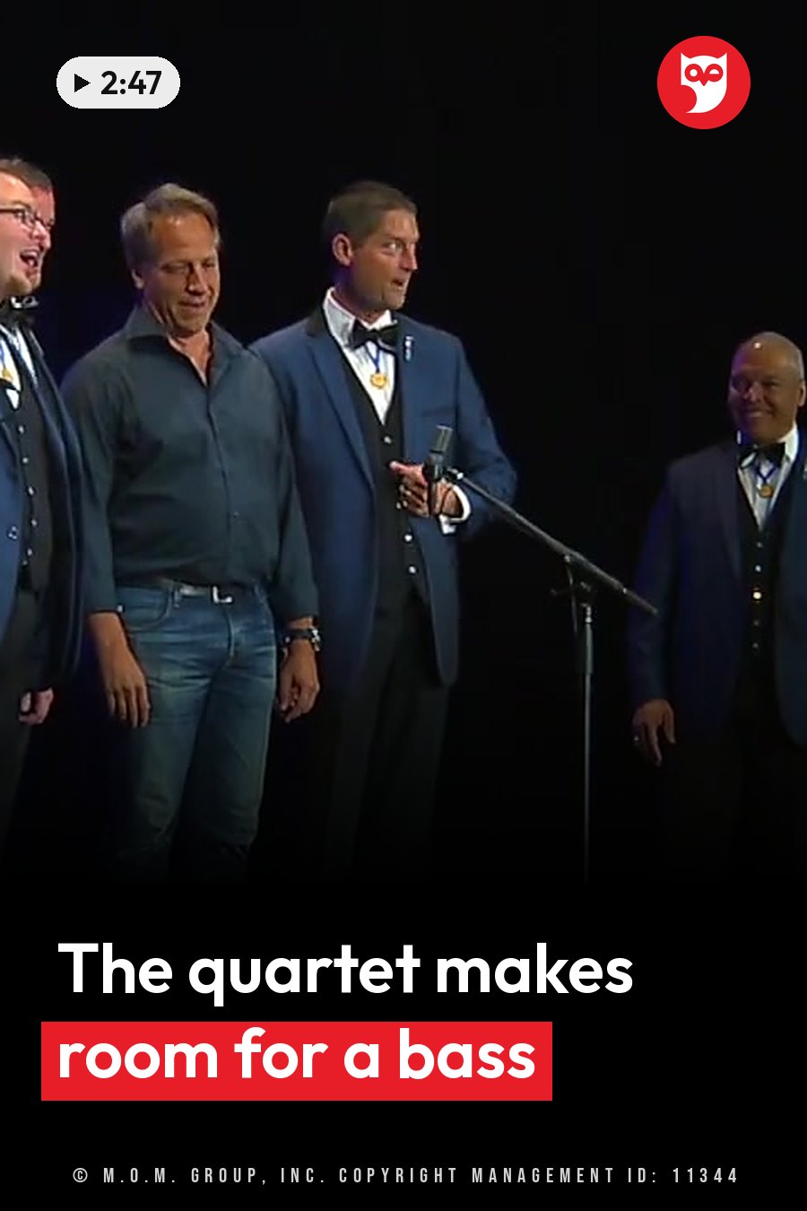 Four men stand on a stage; one speaks into a microphone while the others listen. A headline runs in white letters across the lower third.