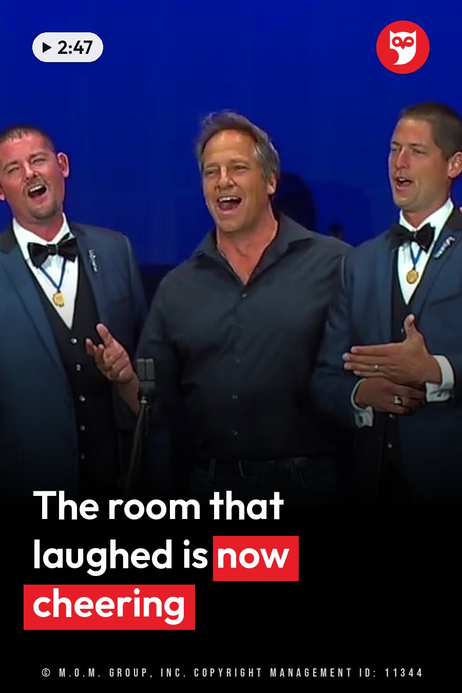 Three men stand on a stage singing into a microphone against a blue background. A headline runs in white letters across the lower third.