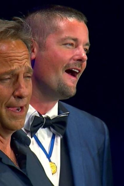 Three middle-aged men dressed formally in an indoor event setting; dark blue backdrop, artificial stage lighting.