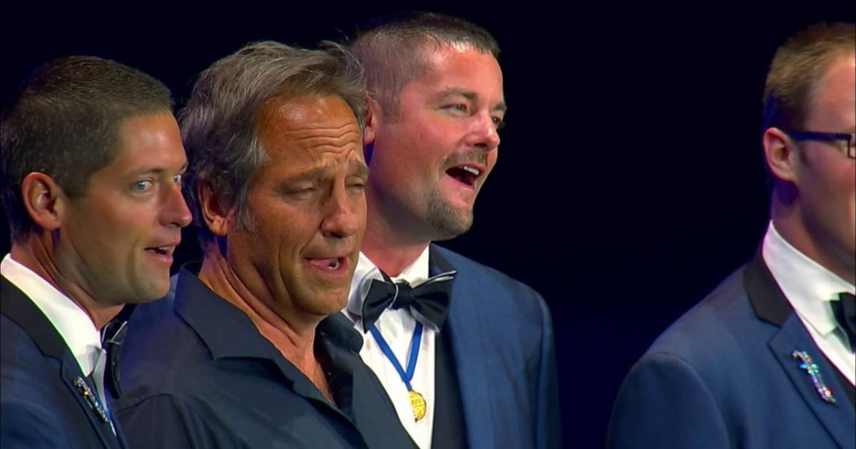 Three middle-aged men dressed formally in an indoor event setting; dark blue backdrop, artificial stage lighting.