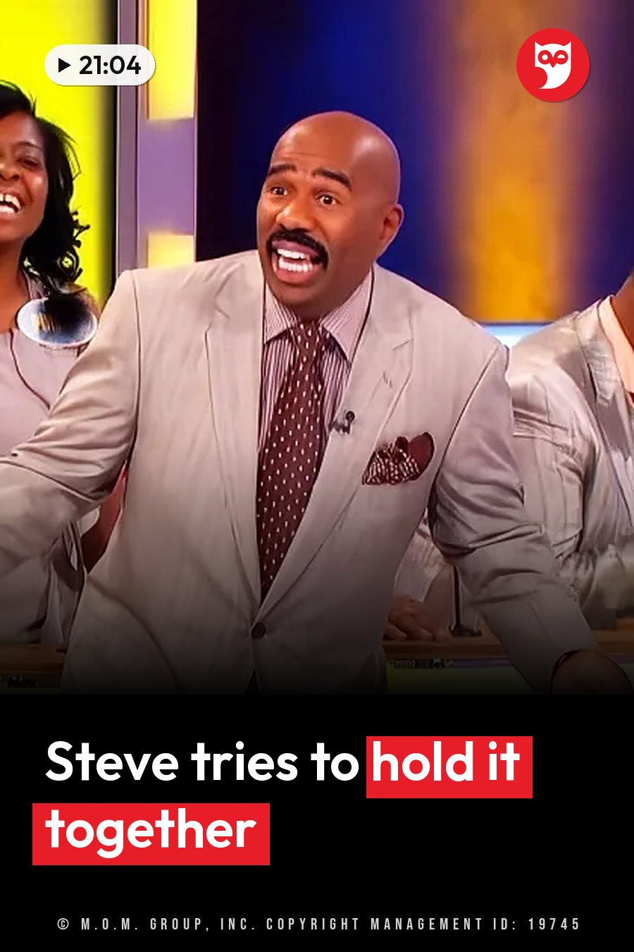 Steve Harvey, a bald man with a mustache in a suit and tie, laughs while seated at a table with other people nearby. A headline runs in white letters across the lower third.