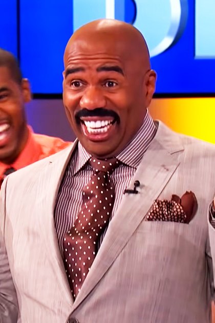 A man in his adult years wearing a light gray suit jacket with a striped shirt and polka dot tie stands on a television show set, surrounded by other adults engaged in dialogue.