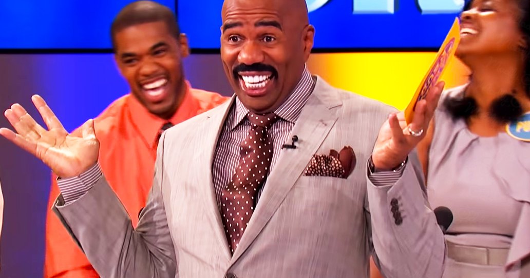 A man in his adult years wearing a light gray suit jacket with a striped shirt and polka dot tie stands on a television show set, surrounded by other adults engaged in dialogue.
