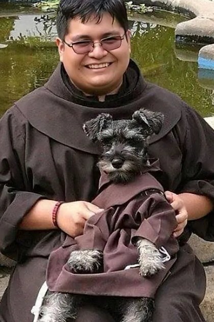A small dog in a monk's robe sits beside a person also dressed as a monk near a pond.