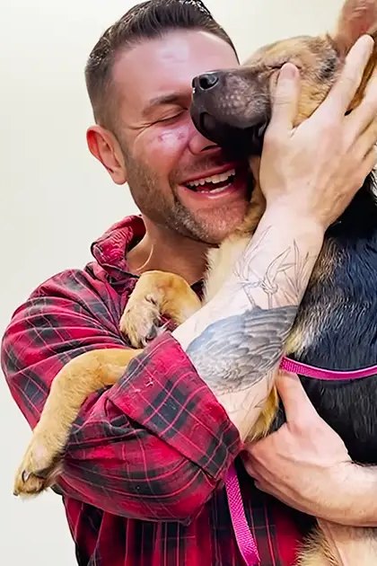 A German Shepherd puppy, likely a young adult, with distinctive coat coloration and pointed ears, held by an adult male in a home setting. The scene is brightly lit, suggesting daytime.
