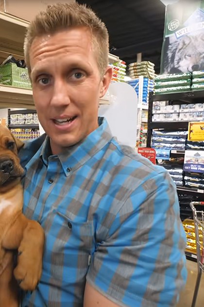 Adult male wearing a blue and gray plaid button-down shirt in a pet store holding a young adult Bloodhound puppy with light brown fur and white chest markings, surrounded by shelves of pet products under even indoor lighting.