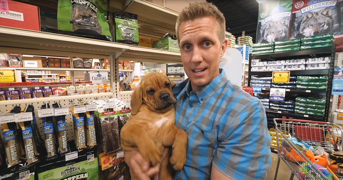 Adult male wearing a blue and gray plaid button-down shirt in a pet store holding a young adult Bloodhound puppy with light brown fur and white chest markings, surrounded by shelves of pet products under even indoor lighting.