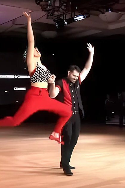A dance couple performs at 'The Open Ballroom Dance Championship' in an indoor ballroom with wooden flooring and overhead lights, with international flags and spectators.