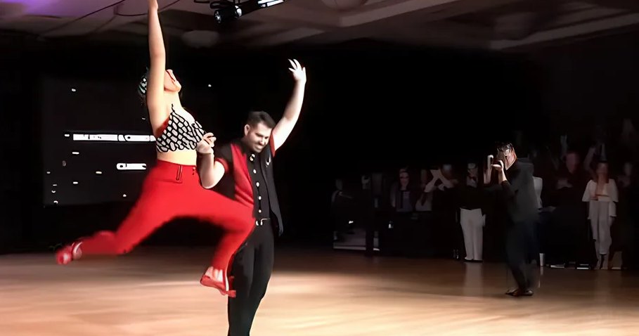 A dance couple performs at 'The Open Ballroom Dance Championship' in an indoor ballroom with wooden flooring and overhead lights, with international flags and spectators.