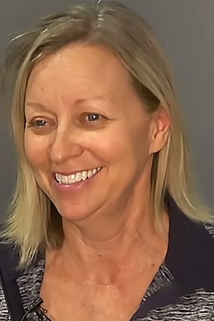 A middle-aged woman stands against a plain, neutral-colored wall indoors. She wears a black and white patterned top with a relaxed fit.
