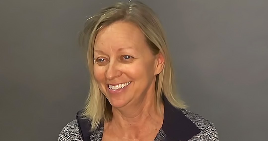 A middle-aged woman stands against a plain, neutral-colored wall indoors. She wears a black and white patterned top with a relaxed fit.