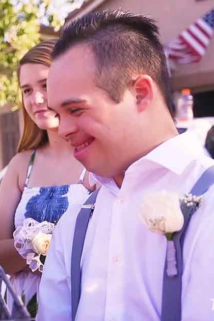 A young adult female news reporter in a black and white patterned dress interviews a young man wearing a white shirt with suspenders and a boutonniere, set against the backdrop of a suburban neighborhood with lush greenery and an American flag visible.