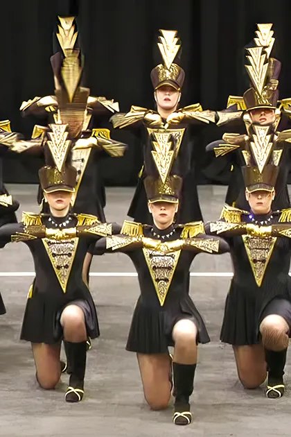 A group performance by young adult female dancers in a dance or marching band setting, wearing form-fitting uniforms with black and gold accents on stage under artificial lighting.