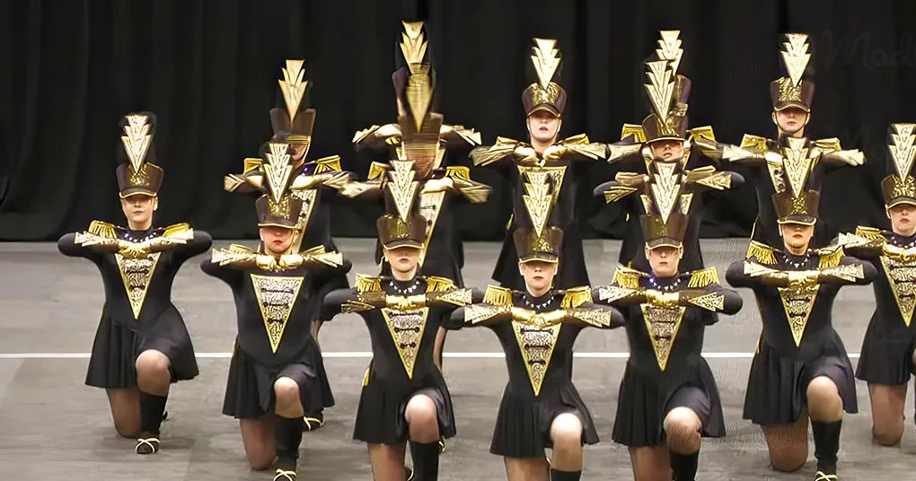 A group performance by young adult female dancers in a dance or marching band setting, wearing form-fitting uniforms with black and gold accents on stage under artificial lighting.