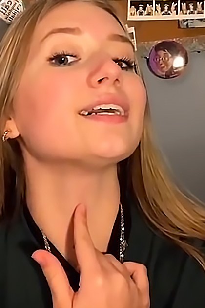 A young adult female wearing a dark green blazer in an indoor setting with artificial lighting, holding a circular decorative object on the wall; another young adult female in a dark green turtleneck sweater and silver necklace, standing near a bed frame under similar lighting conditions.