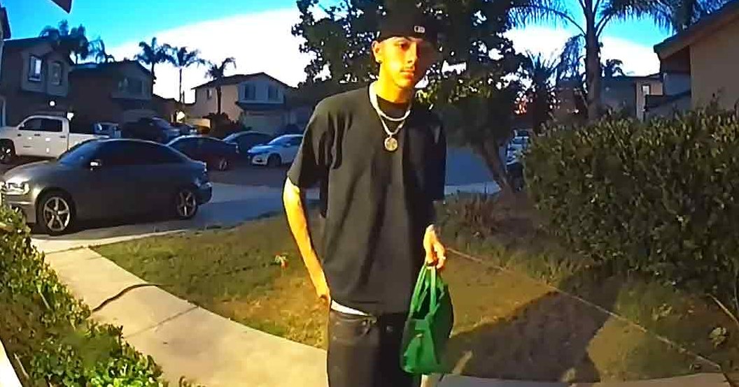 A young adult male in a residential neighborhood, wearing a black t-shirt with dark jeans and a silver chain necklace, holding a green plastic bag on the sidewalk.