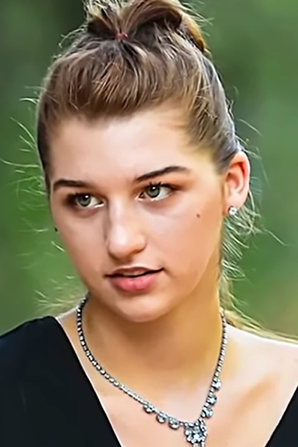 A young adult female wearing a black sleeveless top and small earrings, surrounded by lush greenery under bright natural light.