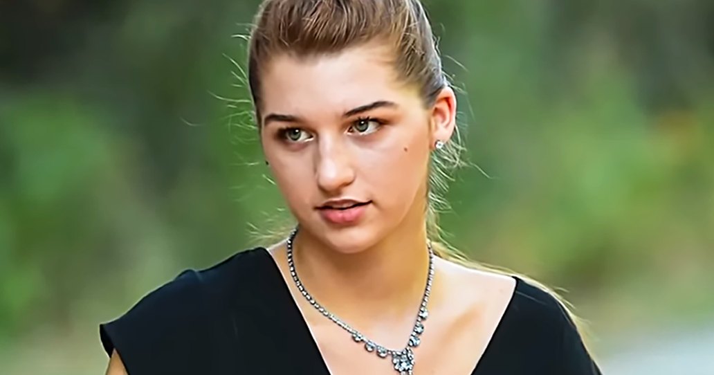 A young adult female wearing a black sleeveless top and small earrings, surrounded by lush greenery under bright natural light.