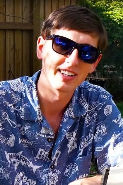 Young adult male wearing a relaxed fit blue Hawaiian shirt with white and yellow patterns, dark sunglasses in an outdoor backyard setting with greenery, wooden fence, and smoke from a fire pit.
