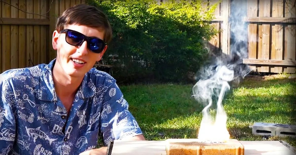 Young adult male wearing a relaxed fit blue Hawaiian shirt with white and yellow patterns, dark sunglasses in an outdoor backyard setting with greenery, wooden fence, and smoke from a fire pit.