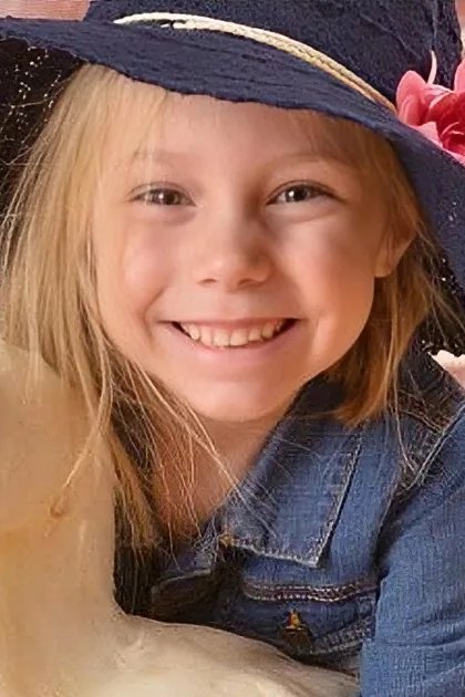 A young child wearing a denim jacket and navy blue hat adorned with a pink flower, standing in front of a brick wall background.