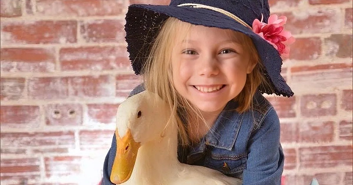 A young child wearing a denim jacket and navy blue hat adorned with a pink flower, standing in front of a brick wall background.