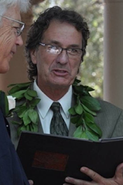A wedding ceremony outdoors with columns in the background. A middle-aged man wearing a light-colored suit jacket, patterned tie, and lei holds what appears to be a book or document. An older adult female wears glasses, a white lace dress with floral embellishments, and a pearl necklace.