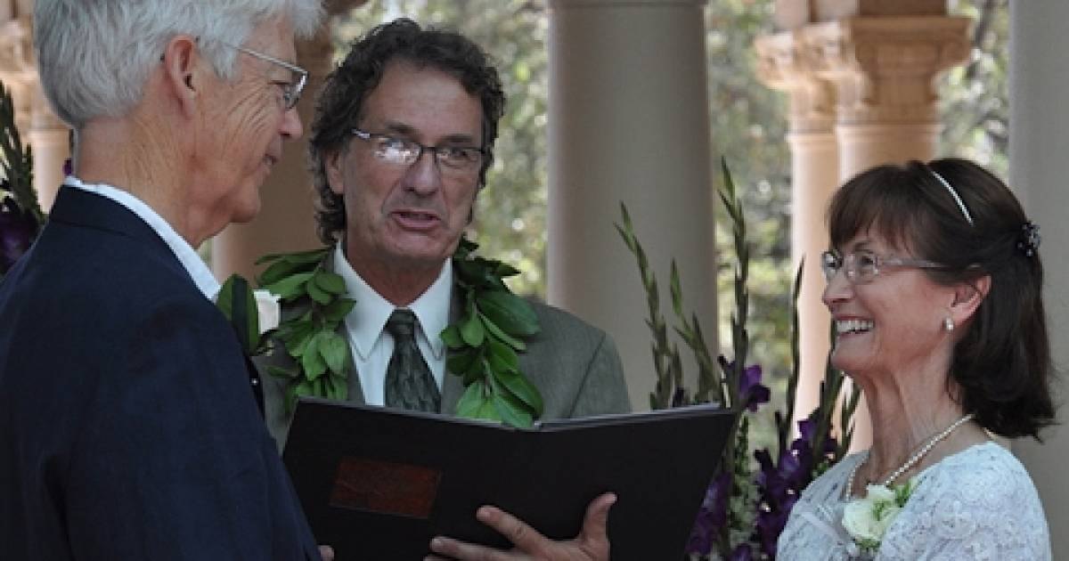 A wedding ceremony outdoors with columns in the background. A middle-aged man wearing a light-colored suit jacket, patterned tie, and lei holds what appears to be a book or document. An older adult female wears glasses, a white lace dress with floral embellishments, and a pearl necklace.