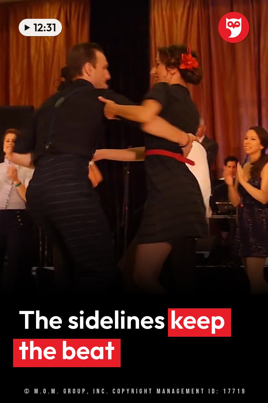 Two people dance on a stage while a group of people stands nearby, watching and clapping. A headline runs in white letters across the lower third.