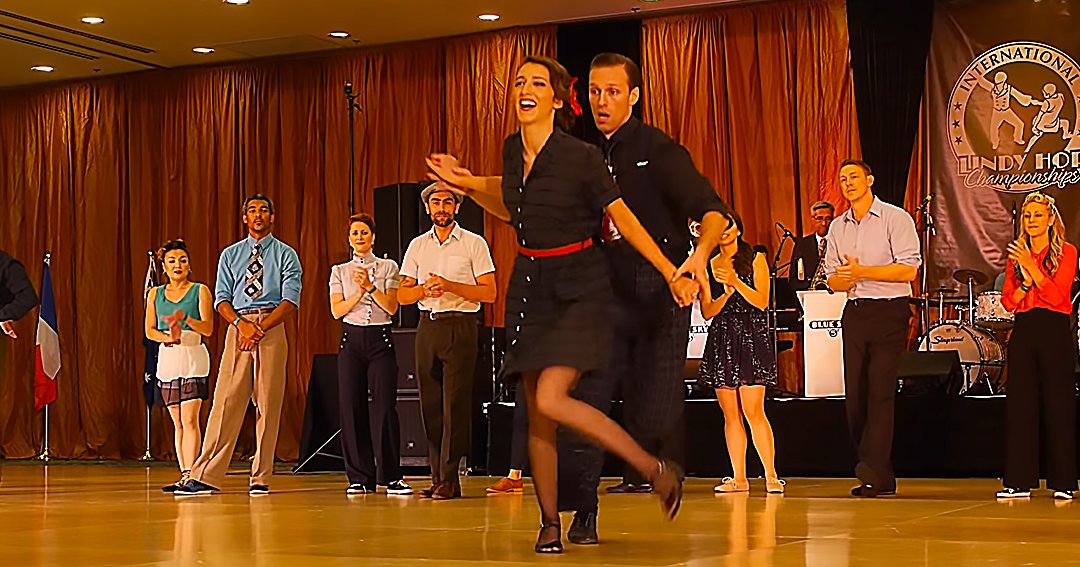 A couple dancing swing in front of an audience at the International Lindy Hop Championships 2021, with a banner reading 'International Lindy Hop Championships,' curtains, and microphones visible.