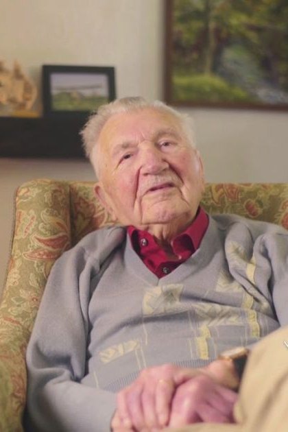 An older adult male wearing a light gray sweater vest over a red collared shirt stands in an indoor living room setting with floral-patterned furniture, artwork on the walls, and soft lighting.