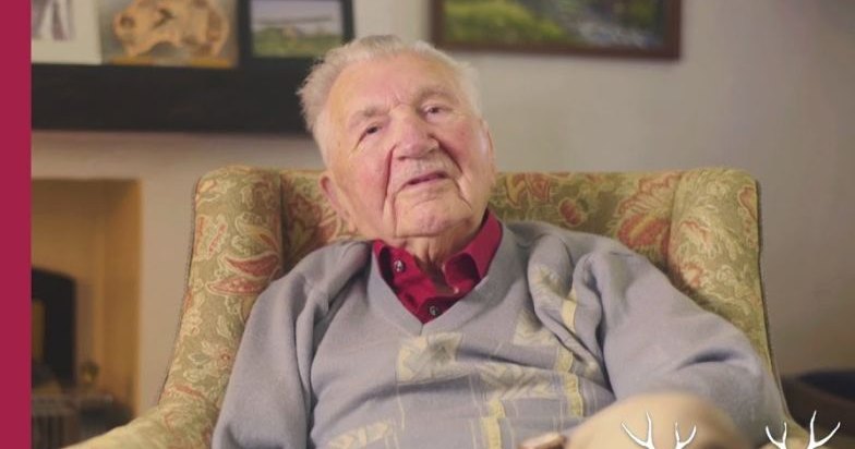 An older adult male wearing a light gray sweater vest over a red collared shirt stands in an indoor living room setting with floral-patterned furniture, artwork on the walls, and soft lighting.