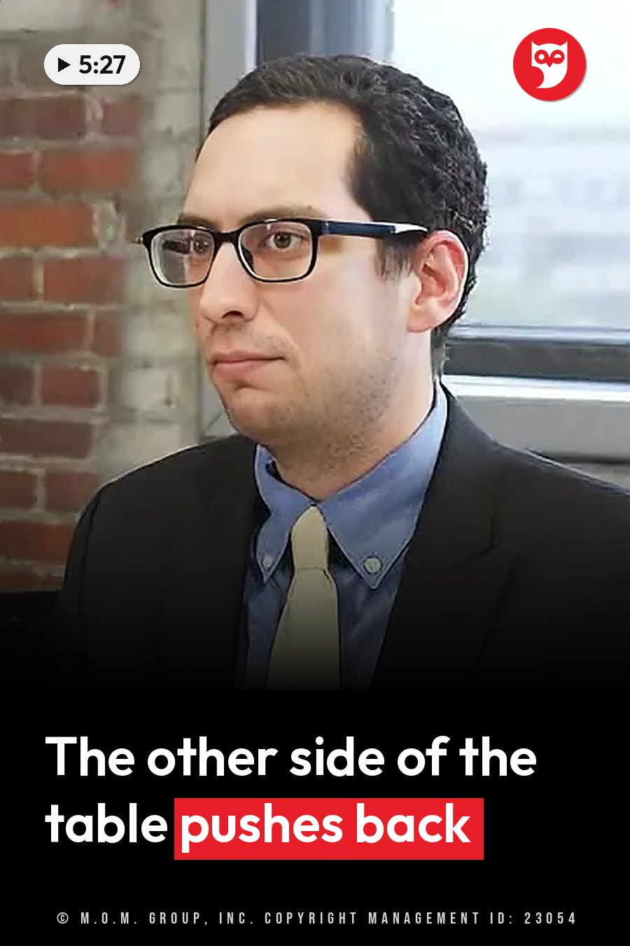 A man with glasses in a suit and tie sits indoors near a brick wall and a window. A headline runs in white letters across the lower third.