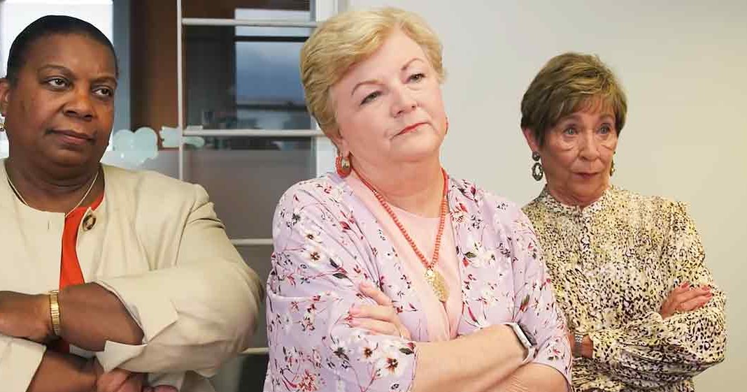 Three women in professional attire, an older adult female left with a beige blazer over red top, middle-aged female center with light pink floral blouse, and older adult right in yellow and white patterned blouse, seated indoors possibly at an office or meeting room setting.