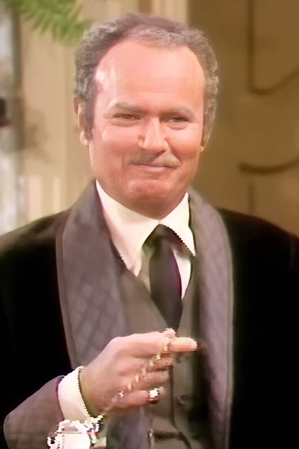 Two men in formal attire holding small objects; dark suits with light shirts and ties, patterned vest visible. Indoor setting with yellow chair and flowers.