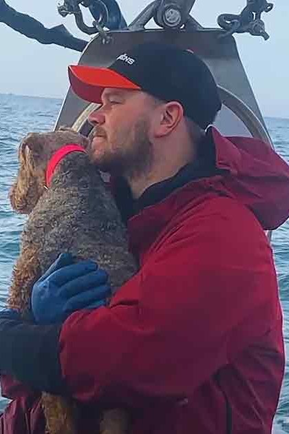 An adult male wearing a red jacket holds a small brown-and-white dog on a boat at sea during colder weather.