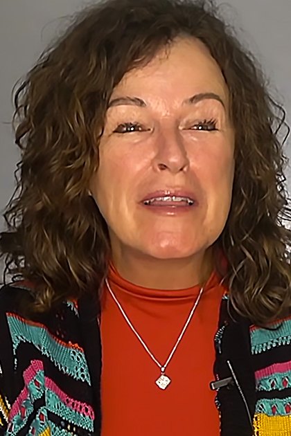 A middle-aged adult female wearing a relaxed fit, black sweater jacket with colorful striped pattern and high neckline indoors against a plain gray background.