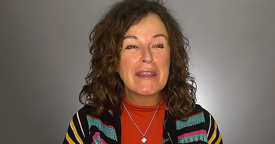 A middle-aged adult female wearing a relaxed fit, black sweater jacket with colorful striped pattern and high neckline indoors against a plain gray background.