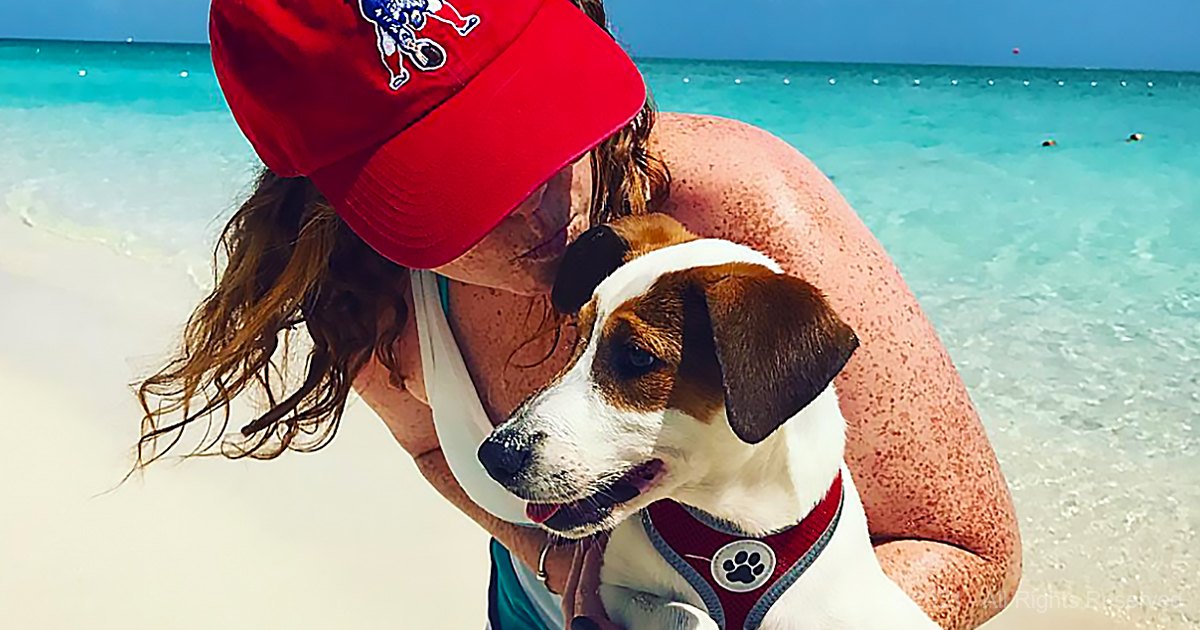 Adult female holding a beagle mix dog at a beach on a bright summer day with clear skies and red, white, and brown colors dominating the scene.