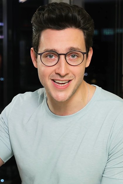 A young adult male wearing a light blue, relaxed t-shirt with round neckline and short sleeves is seated indoors near dining furniture including chairs and a vase of flowers on a table, with artificial lighting indicating evening or nighttime.