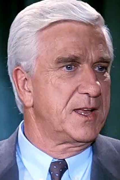 An older adult male in a tailored dark gray suit jacket and light blue collared shirt, set against a dark green curtain backdrop.