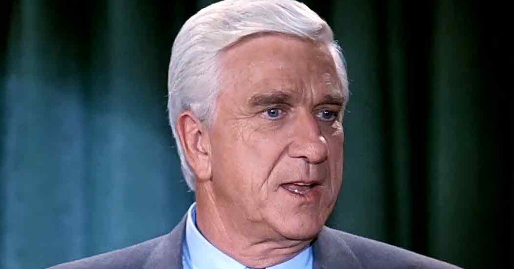 An older adult male in a tailored dark gray suit jacket and light blue collared shirt, set against a dark green curtain backdrop.